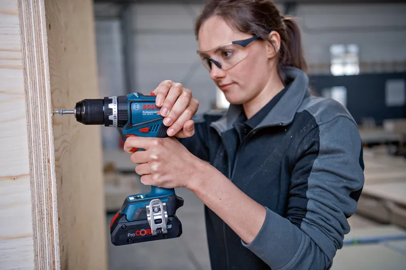 A person wearing safety equipment uses a cordless drill on a wooden panel.