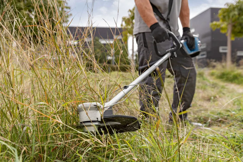 A person wearing safety equipment trims tall grass with a cordless brushcutter.