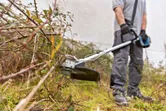 A person wearing safety equipment trims overgrown grass and brush with a cordless brushcutter.