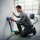 A person wearing safety equipment sands a wall using a random orbit sander with dust extraction.