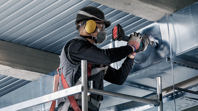 A person wearing safety equipment fastens a metal duct while standing on scaffolding.