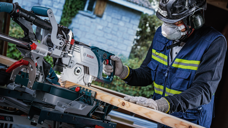 A person wearing safety equipment operates a miter saw to cut a wooden board.