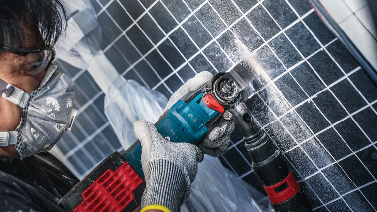 A person wearing safety equipment drills into a tiled wall creating dust.