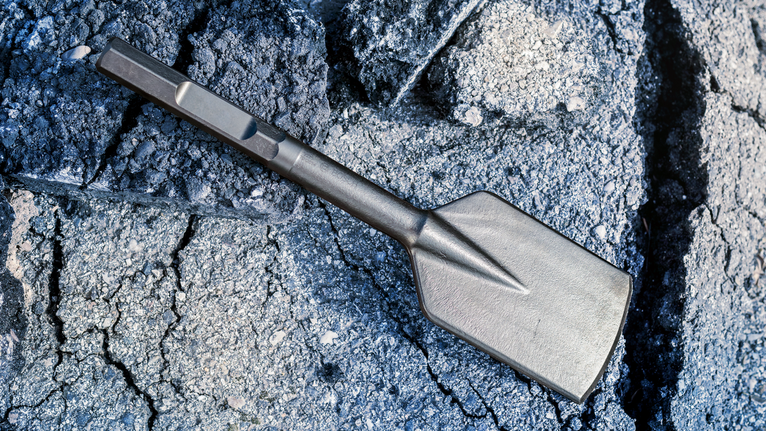 Wide flat chisel rests on cracked concrete surface.
