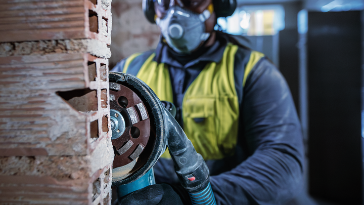 A person wearing safety equipment cuts into a brick wall using a power grinder.