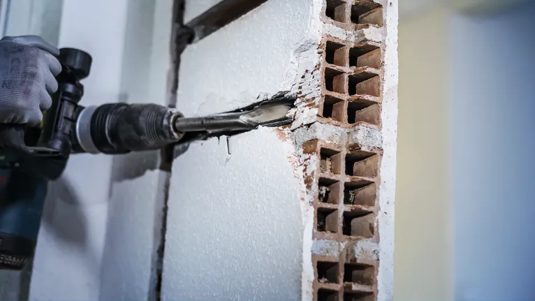 A person wearing safety equipment uses a rotary hammer to chip away a brick wall.