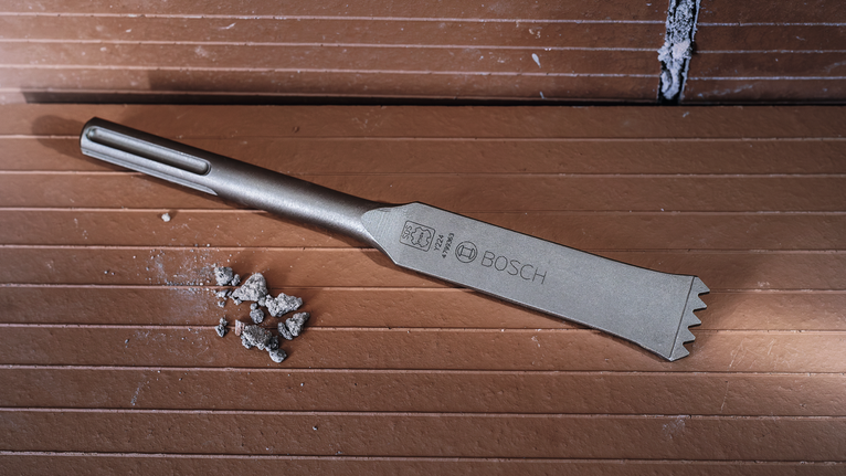 Wide chisel laying on a grooved tiled surface with small pieces of debris nearby.