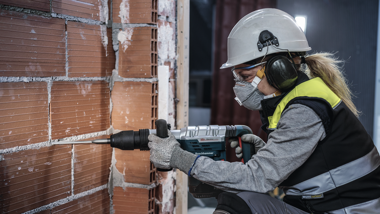 Person wearing safety equipment uses a rotary hammer to chisel into a brick wall.