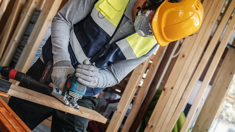 A person wearing safety equipment uses a jigsaw to cut a wooden board.