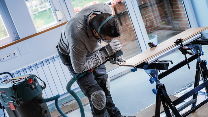 Person wearing safety equipment cuts wood with a power tool on a workbench.
