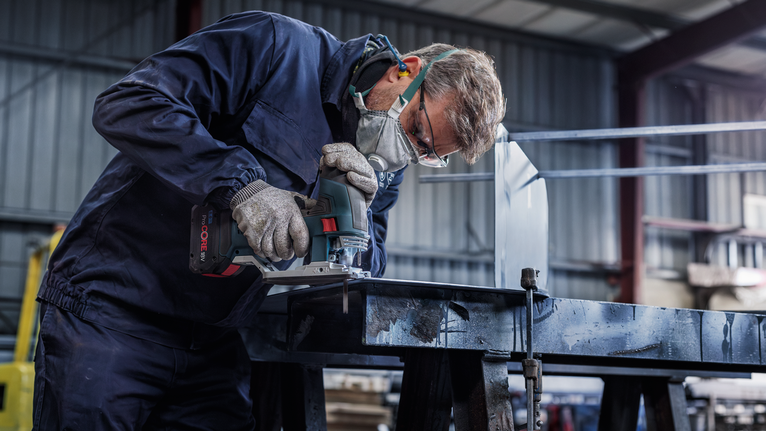A person wearing safety equipment uses a power tool to cut metal in a workshop.