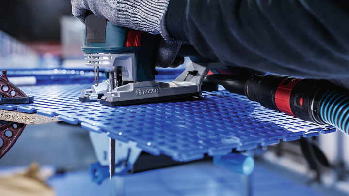 Person wearing safety equipment guides a jigsaw to cut a blue perforated sheet.