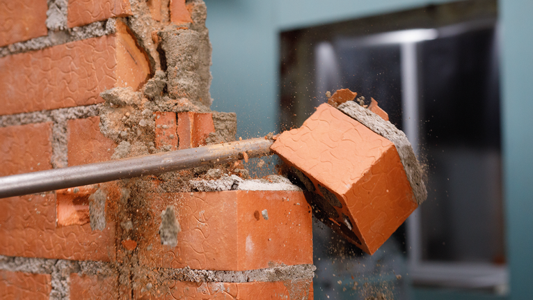 Masonry chisel breaking a brick in a partially demolished wall.