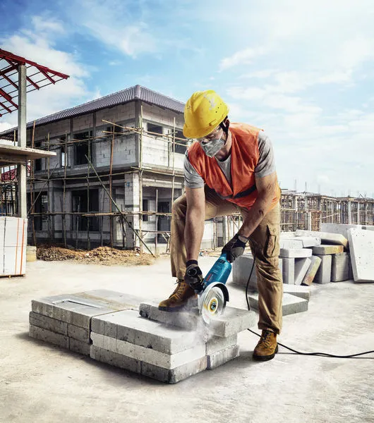 A person wearing safety equipment cuts concrete blocks with an angle grinder at a construction site.