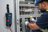 Person wearing safety equipment tests electrical panel connections with a digital multimeter.