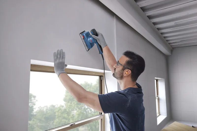 A person wearing safety equipment sands a wall near a window using an orbital sander.