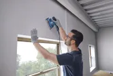 A person wearing safety equipment sands a wall near a window using an orbital sander.