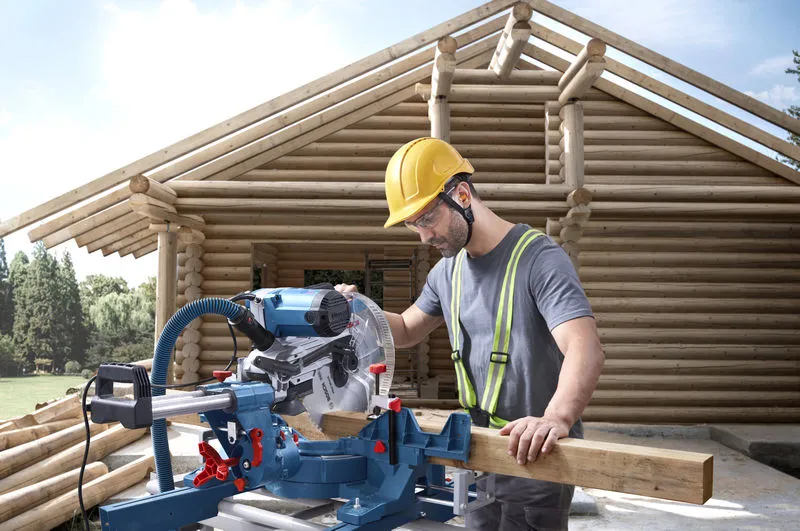 A person wearing safety equipment cuts wood with a mitre saw at a log house construction site.