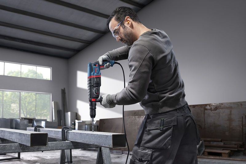 A person wearing safety equipment drills into a steel beam with a rotary hammer.