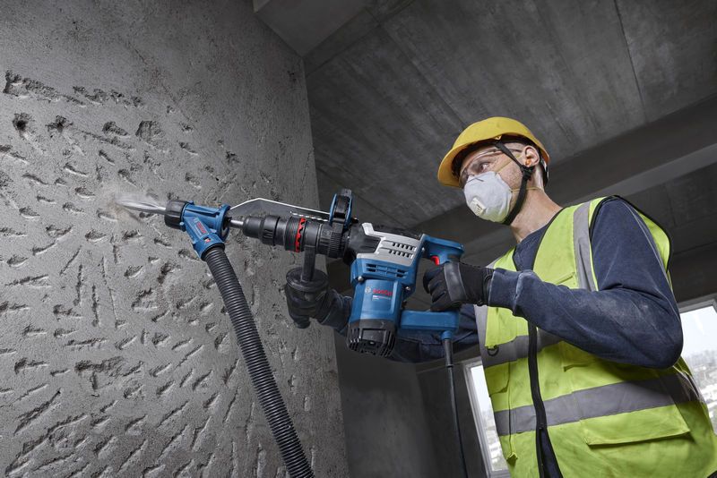 A person wearing safety equipment uses a demolition hammer on a concrete wall.