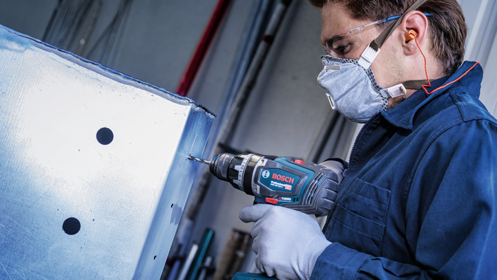 A person wearing safety equipment drills a hole in a metal sheet.