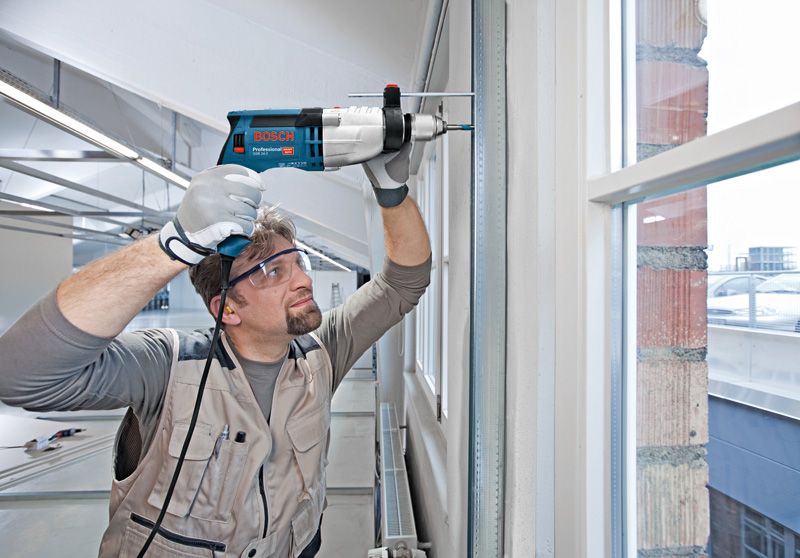 A person wearing safety equipment drills into a wall frame near a window.