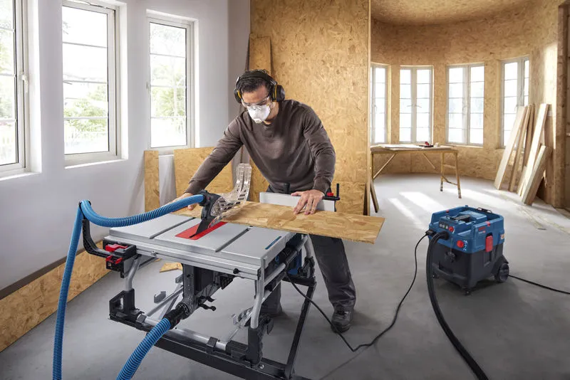 A person wearing safety equipment cuts wood on a table saw in a workshop.
