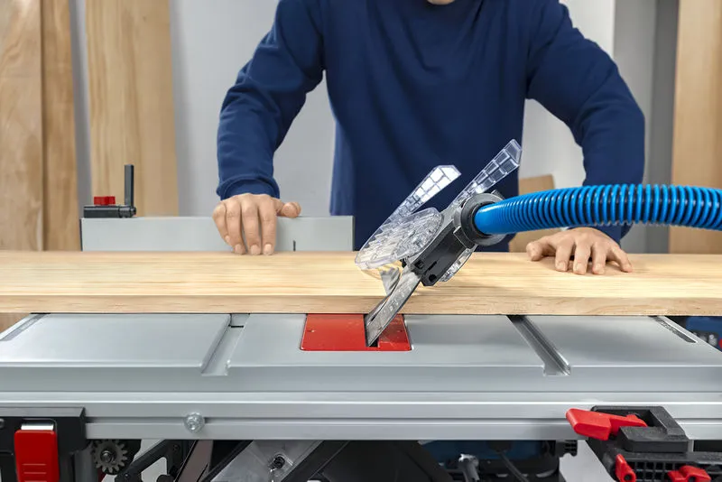 A person guides wood through a table saw with dust extraction hose attached.