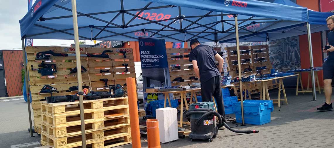 Stół wystawowy Bosch z narzędziami, mężczyzna przy pracy w tle, niebieski namiot.
