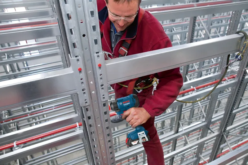 Osoba ubrana w sprzęt bezpieczeństwa obsługuje wiertarkę elektryczną na metalowej belce w magazynie.