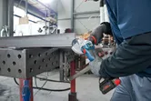 Osoba ubrana w sprzęt ochronny szlifuje belkę metalową za pomocą bezprzewodowej szlifierki kątowej.