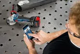 Osoba używająca aplikacji na smartfonie do sterowania bezprzewodową szlifierką kątową na metalowym stole roboczym.