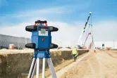 Urządzenie do poziomowania laserowego ustawione na statywie na placu budowy. W pobliżu znajduje się osoba ubrana w sprzęt zabezpieczający.