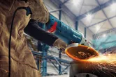 Osoba ubrana w sprzęt ochronny szlifuje metal w warsztacie, co powoduje powstawanie jasnych iskier.