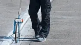 Osoba ubrana w sprzęt bezpieczeństwa mierzy odległość na drodze za pomocą koła pomiarowego.