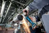 Osoba ubrana w sprzęt ochronny szlifuje metalową rurę za pomocą bezprzewodowej szlifierki kątowej.