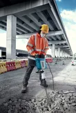Osoba ubrana w sprzęt bezpieczeństwa rozbija beton dużym młotem elektrycznym na zewnątrz budynku.