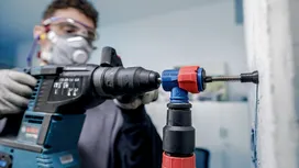 Pracownik ubrany w sprzęt ochronny wierci w ścianie za pomocą elektronarzędzia i przystawki do odsysania pyłu.