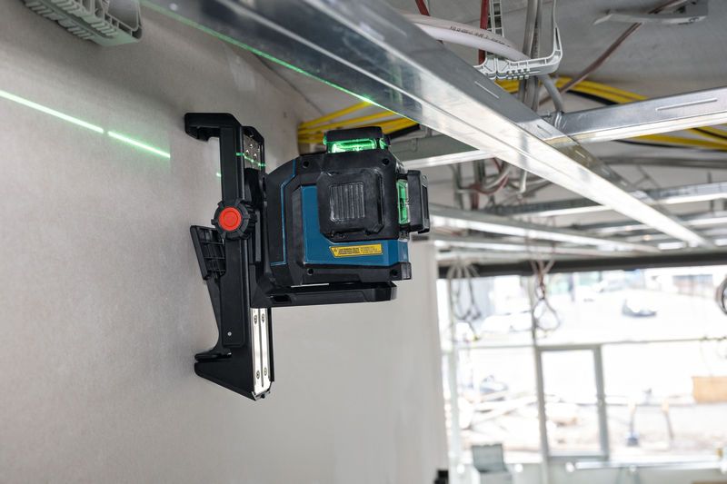 Urządzenie do poziomowania laserowego zamontowane na ścianie, rzucające zieloną linię laserową na plac budowy.
