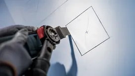 Osoba ubrana w sprzęt ochronny używa elektronarzędzia do wycięcia kwadratu w płycie gipsowo-kartonowej.
