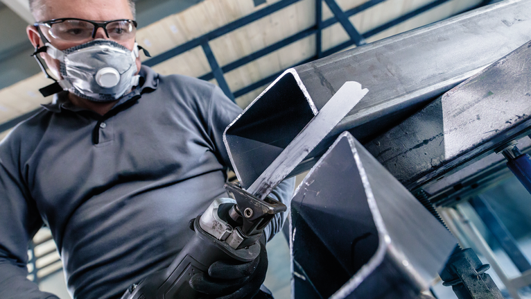 Osoba ubrana w sprzęt ochronny przecina metalową belkę piłą szablastą.