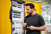 Osoba testuje podzespół w otwartym panelu elektrycznym za pomocą multimetru cyfrowego.