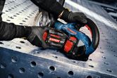 Osoba ubrana w sprzęt ochronny obsługuje bezprzewodową szlifierkę kątową na perforowanej powierzchni metalowej.