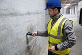 Osoba ubrana w sprzęt bezpieczeństwa sprawdza wilgotność ściany betonowej za pomocą miernika wilgotności materiału.