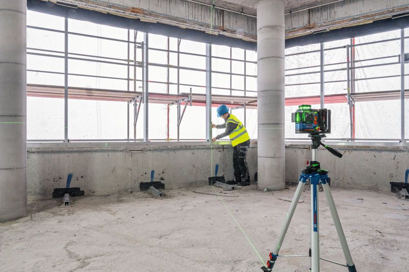 Osoba ubrana w sprzęt bezpieczeństwa wyrównuje ścianę za pomocą niwelatora laserowego umieszczonego na statywie.