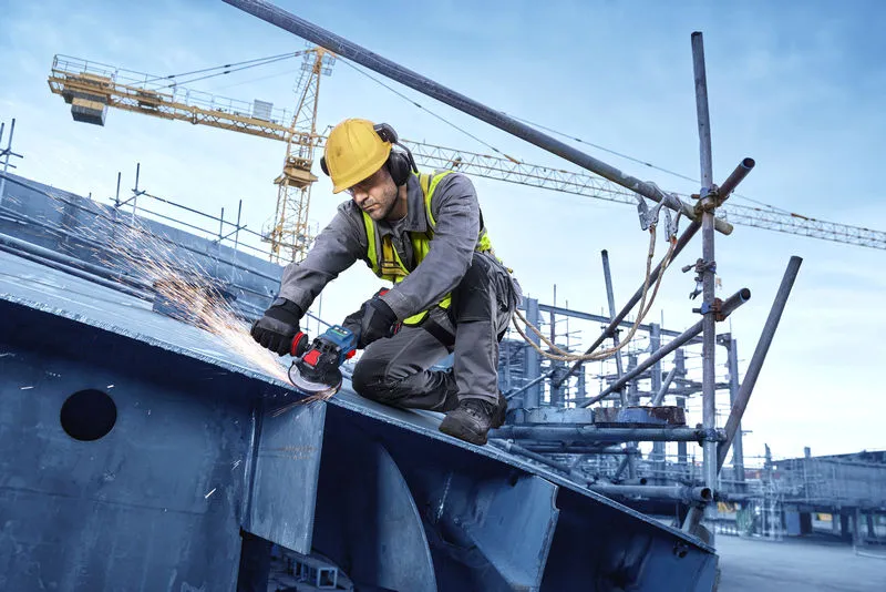 Osoba ubrana w sprzęt ochronny szlifuje belki metalowe za pomocą bezprzewodowej szlifierki kątowej.