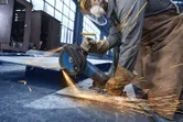 Osoba ubrana w sprzęt ochronny szlifuje metal szlifierką kątową, wytwarzając iskry.