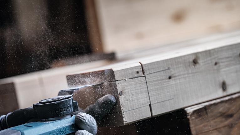 Osoba ubrana w sprzęt ochronny szlifuje drewnianą deskę za pomocą elektronarzędzia.