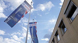 Bandeiras da Bosch flutuam ao vento em frente a um edifício moderno sob céu azul.