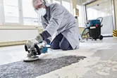 Uma pessoa que utilize equipamento de segurança lixa o pavimento de betão com uma lixadora de betão.